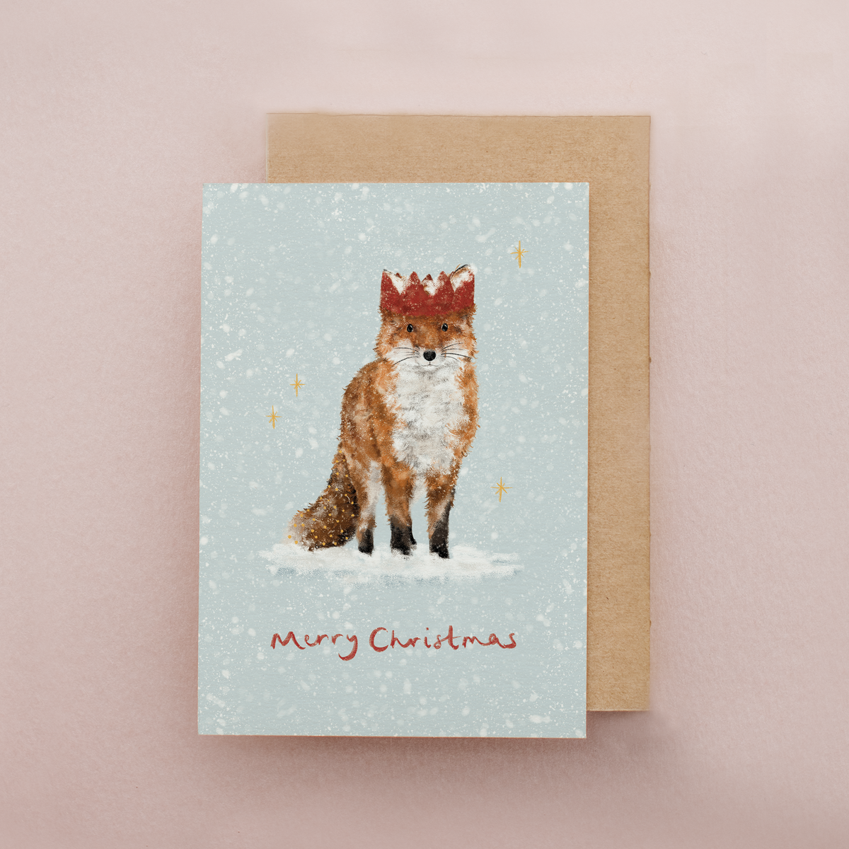 A Christmas greeting card featuring an illustration of a fox wearing a red crown in the snow, with 'Merry Christmas' written below, shown with a brown kraft envelope on a soft pink background.
