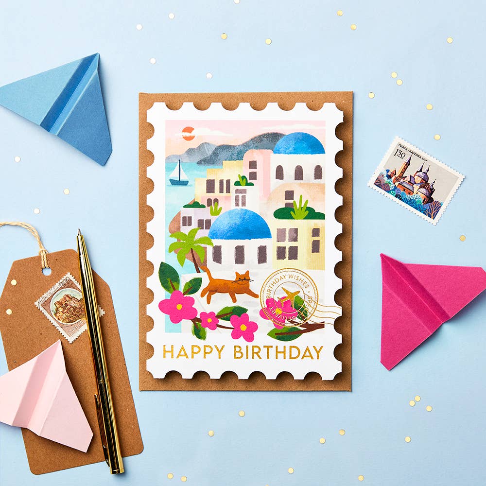 Birthday card with a scenic design of Santorini on a blue tabletop styled with paper planes, a stamp and a luggage tag.