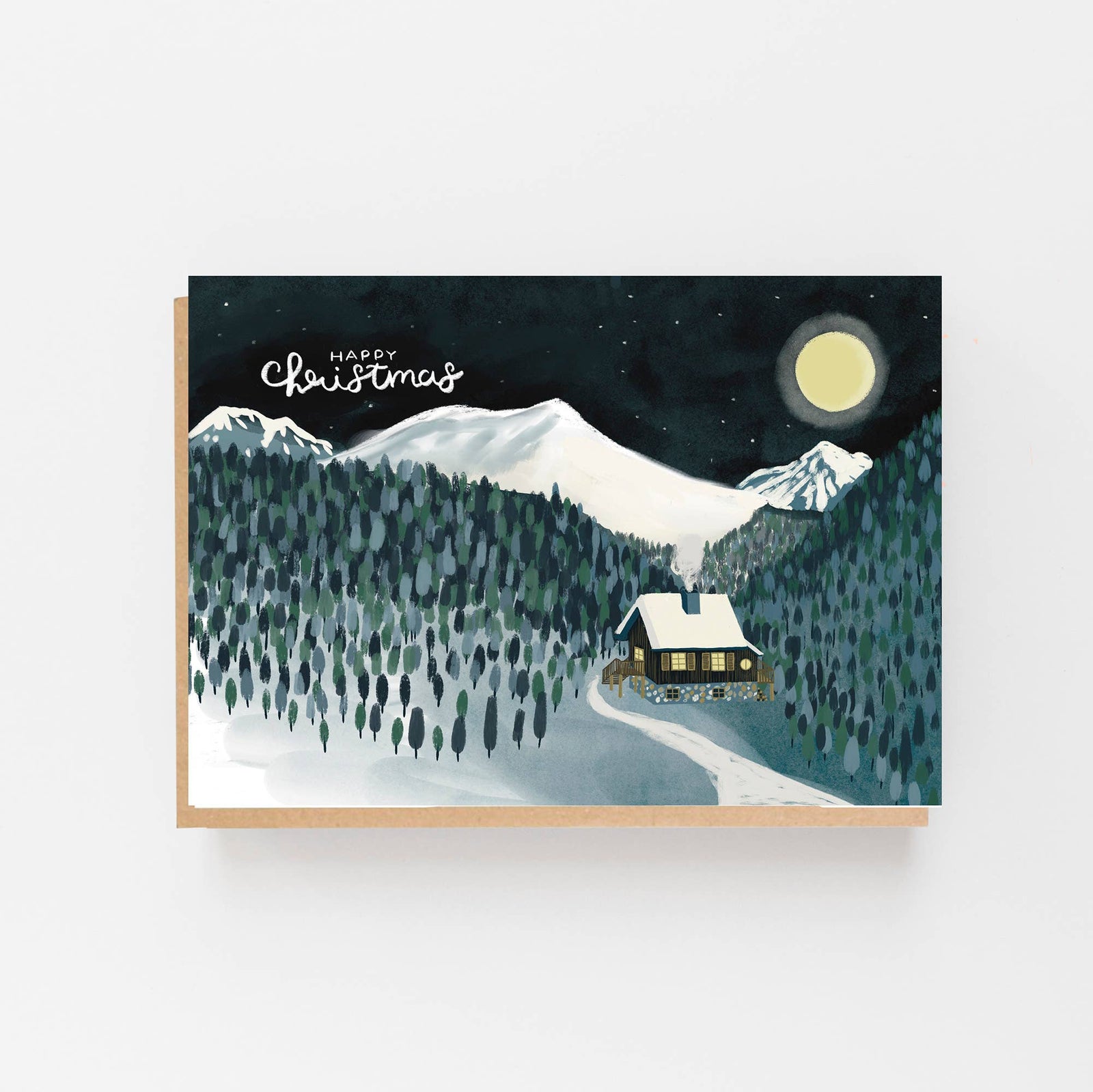 An A6 Christmas greeting card with a kraft paper envelope, featuring an illustration of a snowy mountain landscape at night with a cabin, trees, stars, and a full moon. The card is displayed against a clean white background.