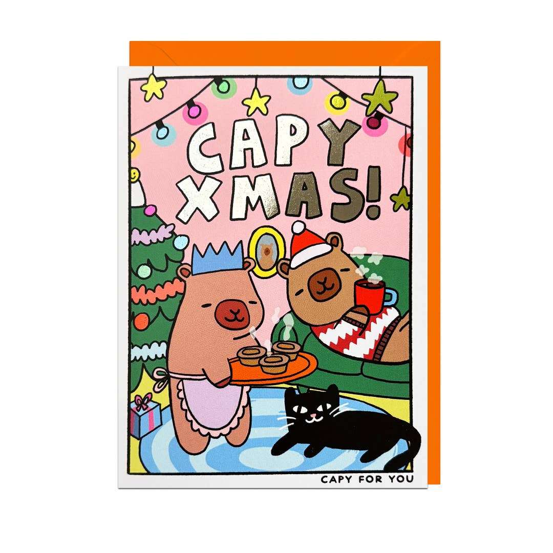 A Christmas-themed greeting card with an orange envelope behind it. The card features a cartoon illustration of two capybaras enjoying mince pies and hot chocolate, a black cat, a Christmas tree, and the text 'CAPY XMAS!'.