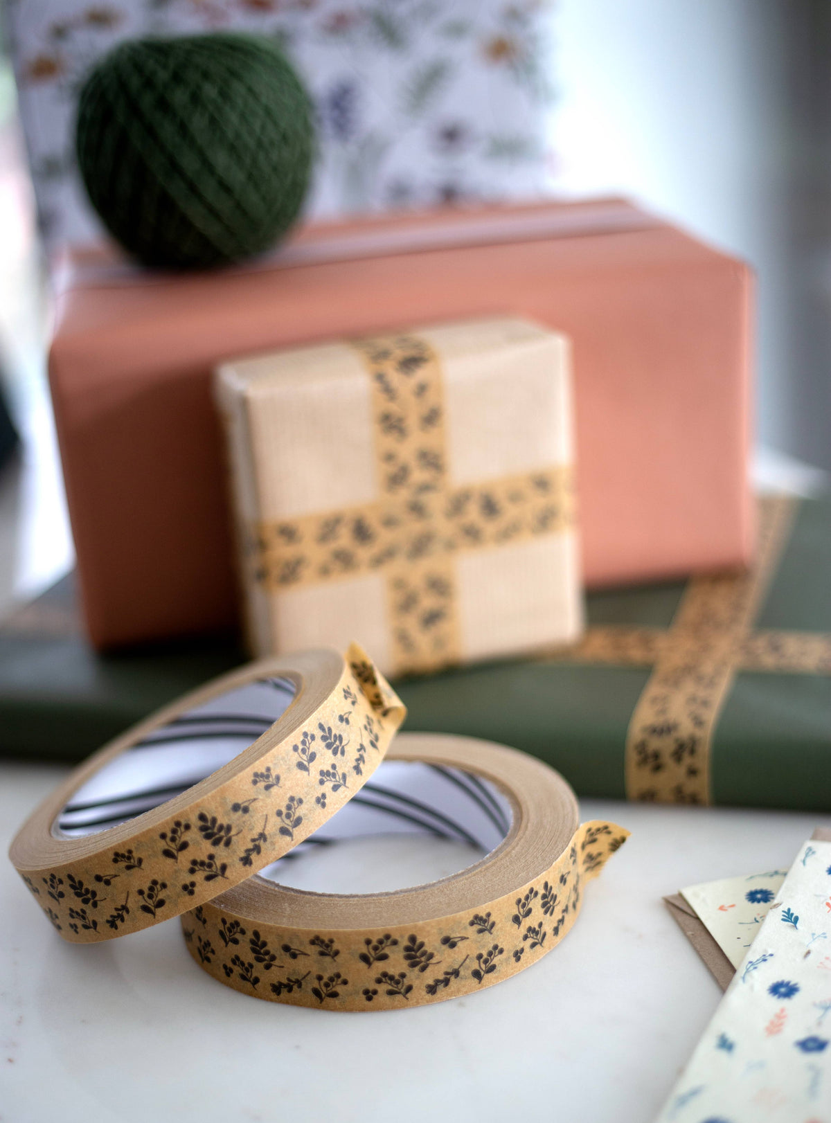 Two rolls of decorative paper tape with a black botanical pattern on a kraft background, displayed in the foreground with wrapped gift boxes and a green yarn ball in the blurred background.