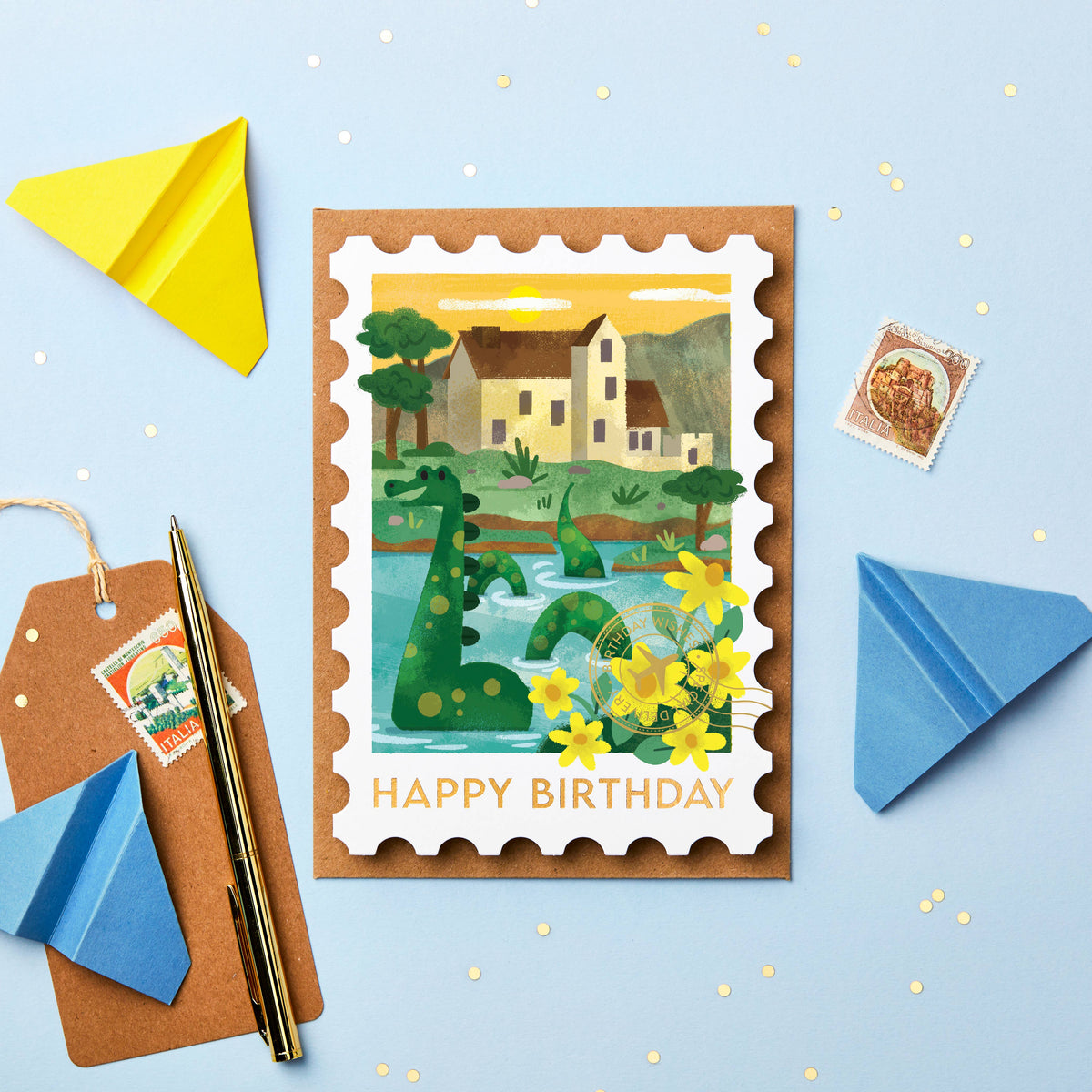 A postage stamp-style birthday card with a stamp border and the words “HAPPY BIRTHDAY” at the bottom, featuring an illustrated Loch Ness Monster in a landscape with flowers. The card is styled on a light blue background with paper airplanes, a pen, a gift tag, and a small postage stamp around it.