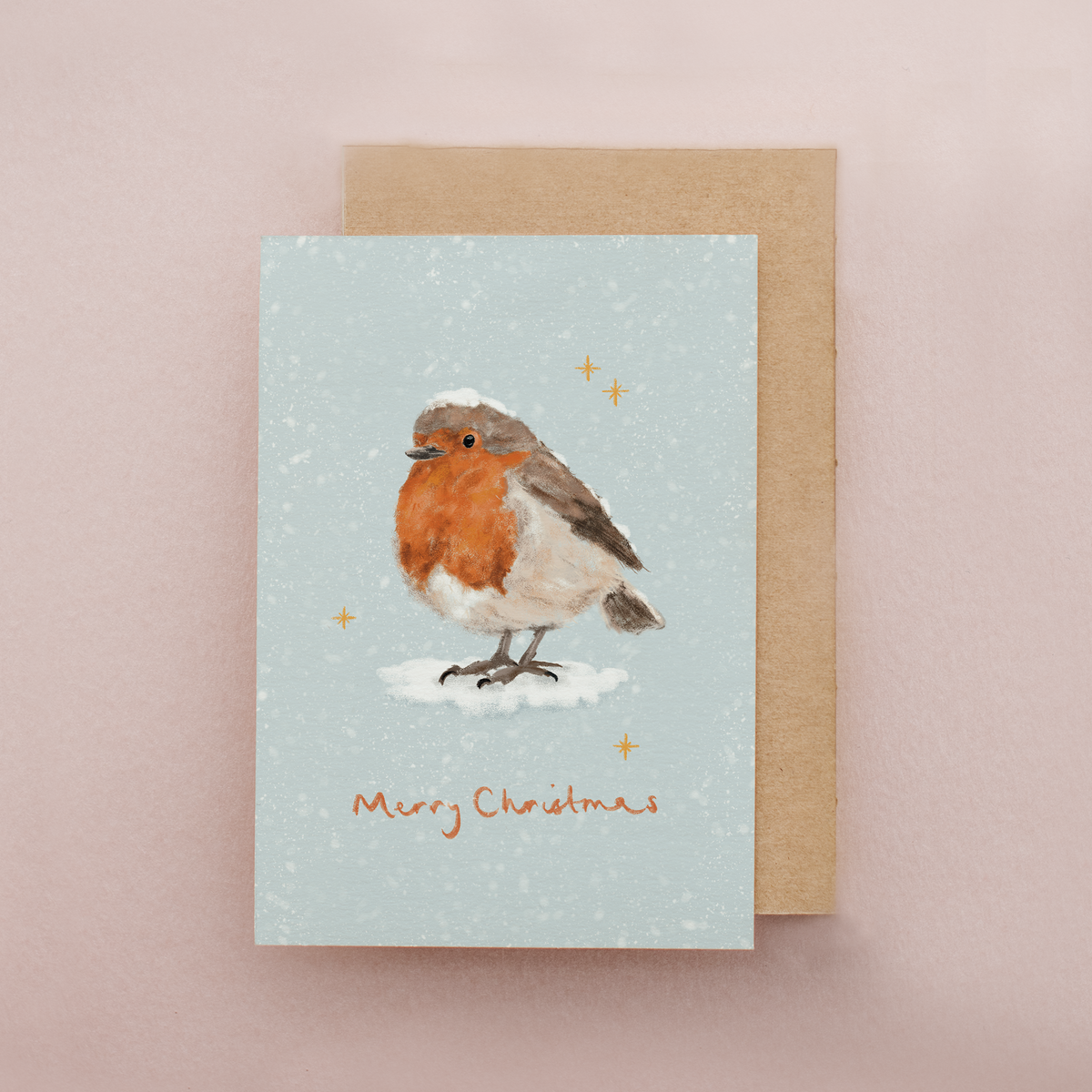 A Christmas greeting card featuring a winter robin standing on snow with small gold stars and the text 'Merry Christmas' at the bottom, shown with a brown kraft envelope on a soft pink background.
