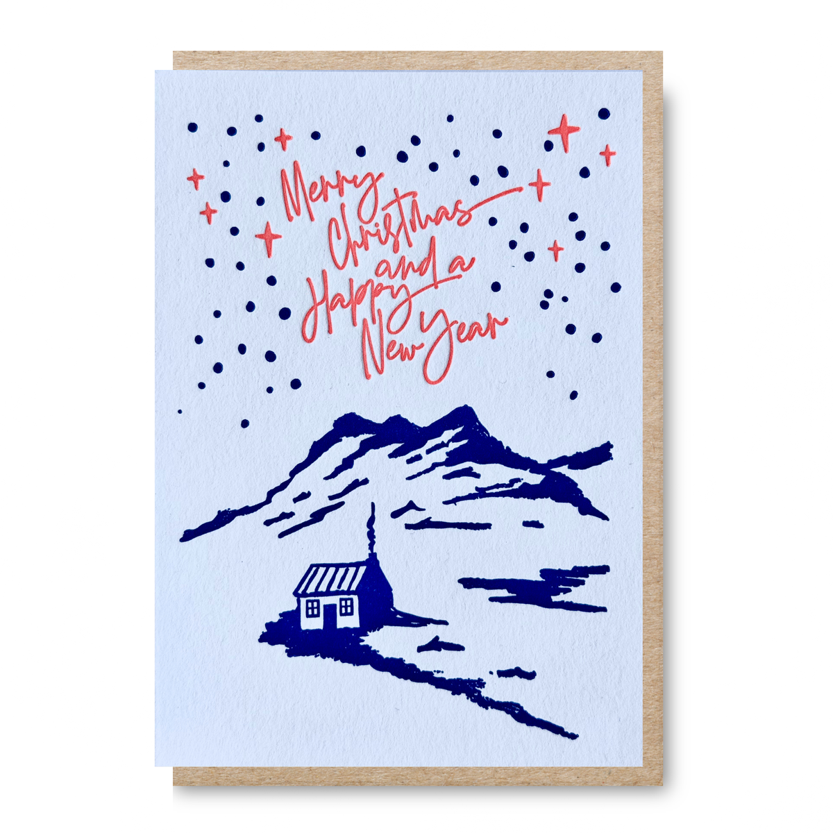 An A6 letterpress printed Christmas greeting card with a kraft envelope, featuring a white background, navy blue mountain and cabin illustration, coral-red script text reading 'Merry Christmas and a Happy New Year,' and scattered navy stars.