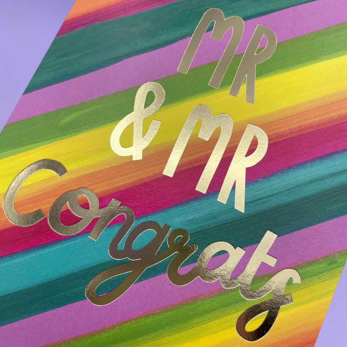 A colourful rainbow stripe greetings card to celebrate a wedding. The rainbow stripes are diagonal across the card and the writing in the centre of the card is big gold foil stating 'Mr & Mr Congrats'.