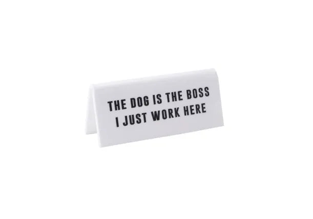 A small white acrylic sign that has in big black capital letters on it 'THE DOG IS THE BOSS I JUST WORK HERE'.