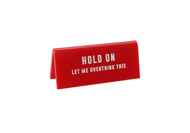 A small red acrylic sign that has in big white capital letters on it 'HOLD ON LET ME OVERTHINK THIS'.
