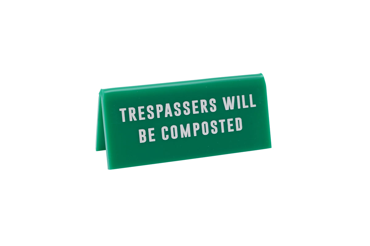 A small GREEN acrylic sign that has in big white capital letters on it 'TRESSPASSERS WILL BE COMPOSTED'.