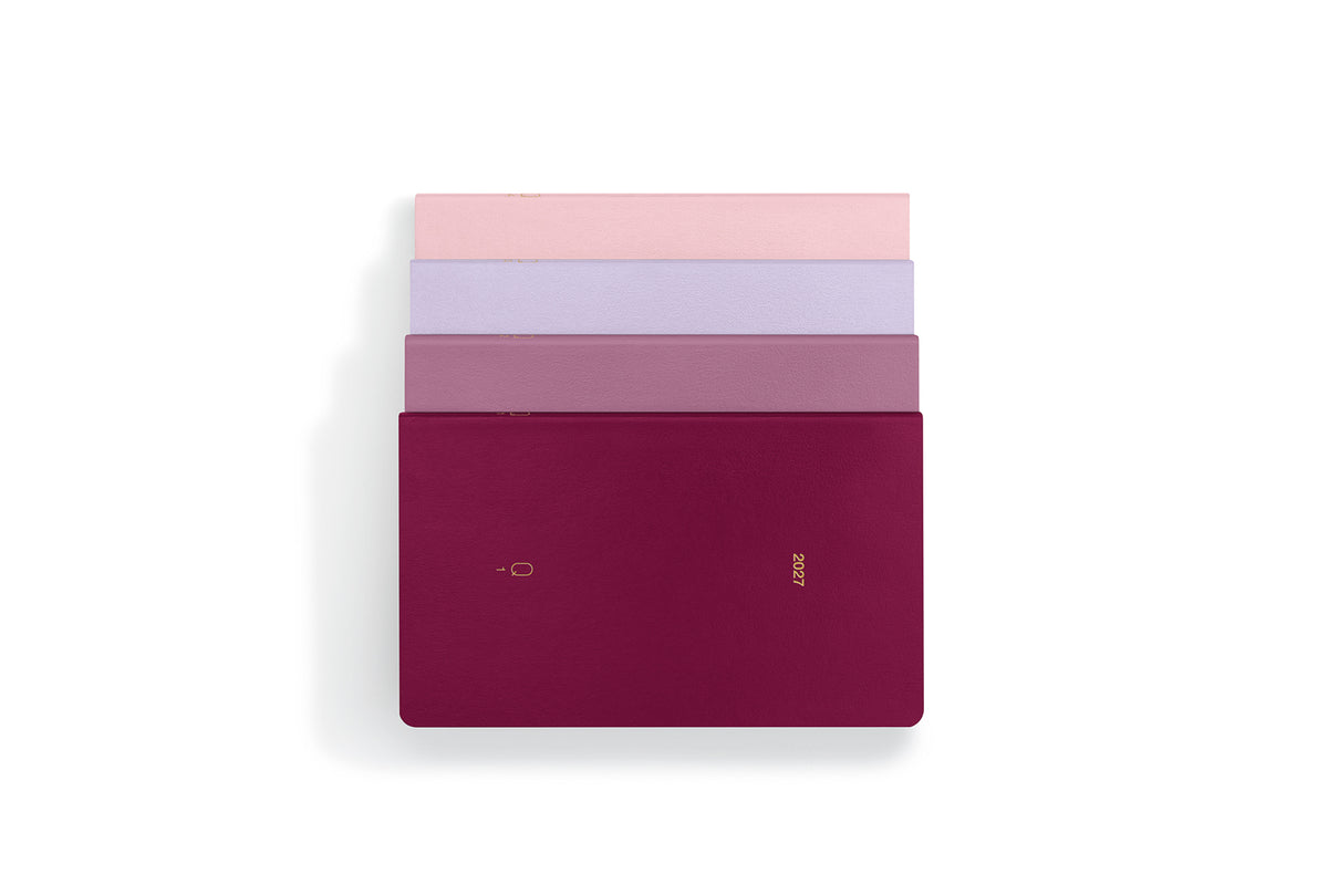 Four slim rectangular notebooks stacked vertically on a white background, with the top three fanned out slightly. The bottom notebook is a deep burgundy/magenta colour with a small gold logo, while the others show pink, lavender, and mauve tones.