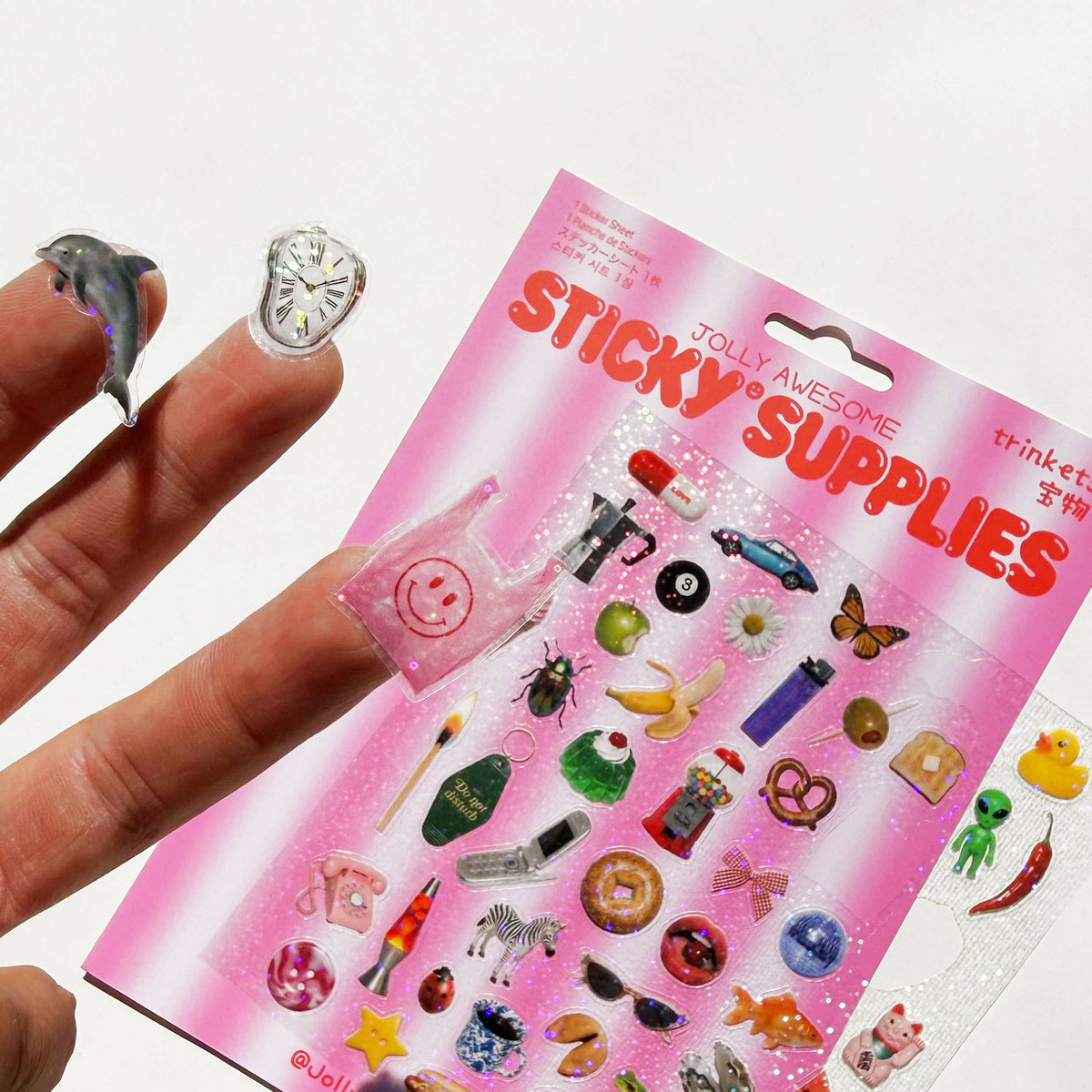 A hand holds 3 small stickers—a dolphin, a plastic carrier bag and a clock—next to a pink package of 'Jolly Awesome Sticky Supplies' trinket stickers.