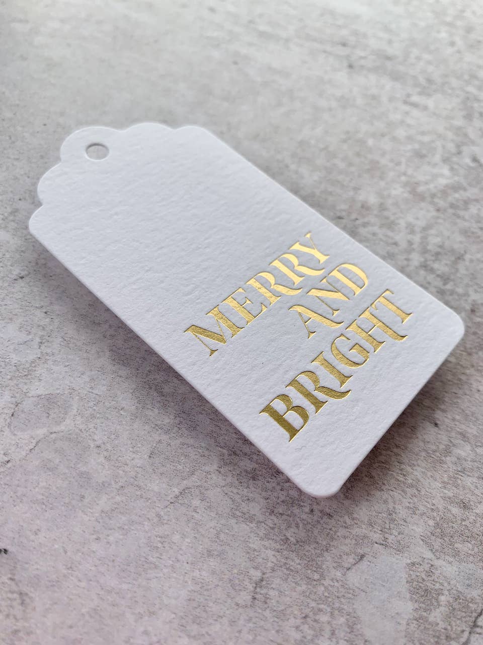 A white textured paper gift tag with scalloped edges and a hanging hole, printed with 'MERRY AND BRIGHT' in metallic gold foil, resting on a light gray textured surface.