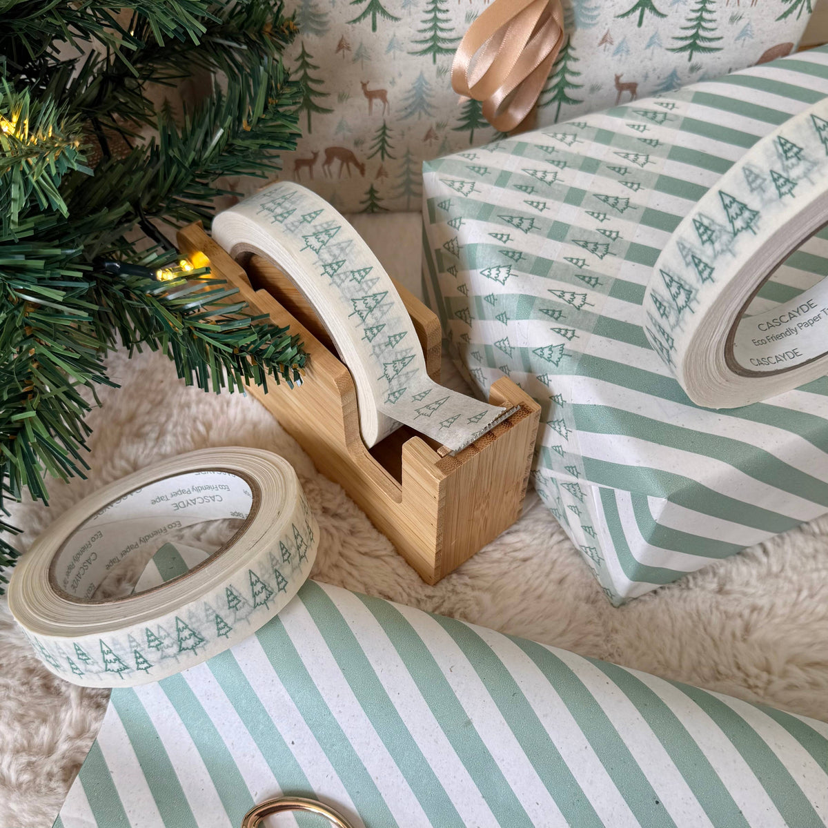 Christmas-themed paper packing tape rolls shown in use, wrapped around gift boxes and displayed on a soft white faux fur surface next to a wooden tape dispenser and a small Christmas tree.