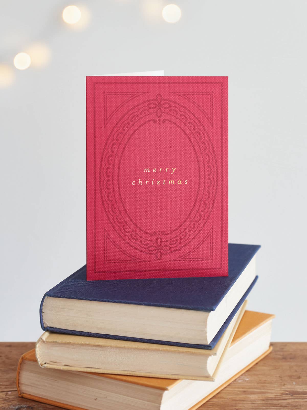 A red Christmas greeting card with an embossed oval design and the words 'merry christmas' in the centre, displayed on top of a stack of books on a wooden surface with soft bokeh lights in the background.
