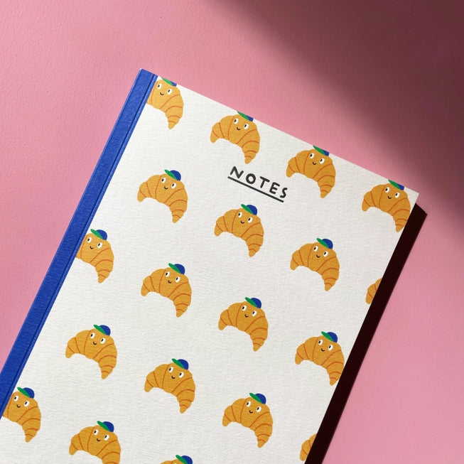 An image of a cream coloured notebook covered in illustrations of croissants with a baseball cap on and face. The notebook has a navy binding and the word 'notes' written in silver foil.