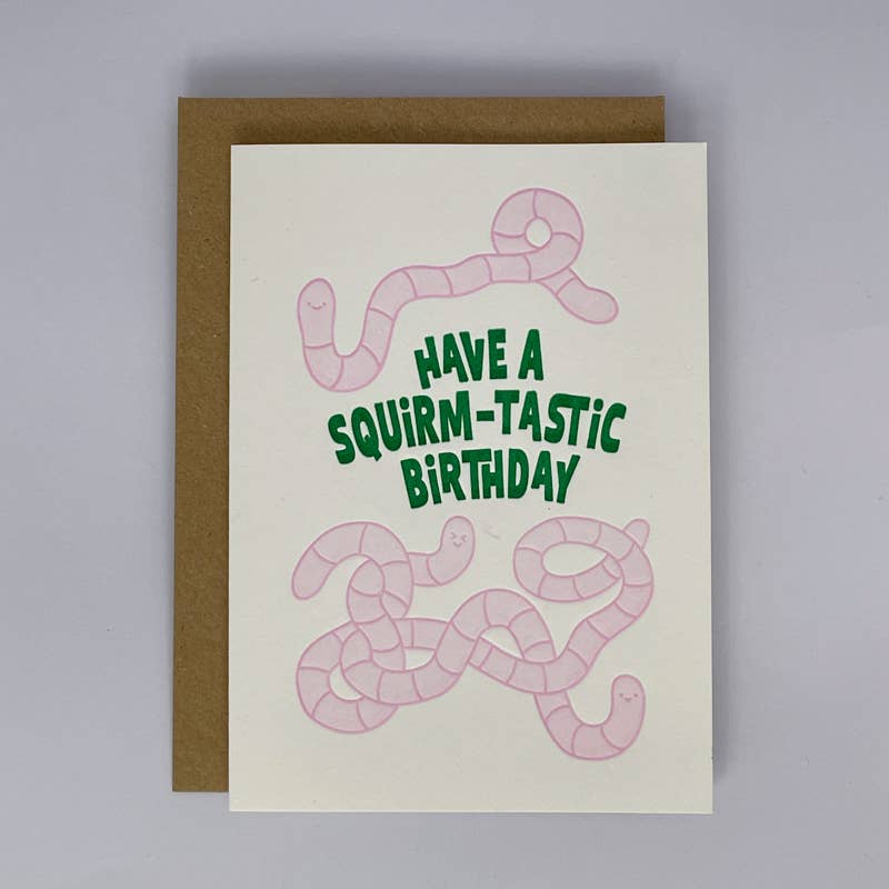 An A6 greeting card with a brown envelope, featuring pink cartoon worms and the text 'HAVE A SQUIRM-TASTIC BIRTHDAY' in green on an off-white card, photographed on a light gray background.