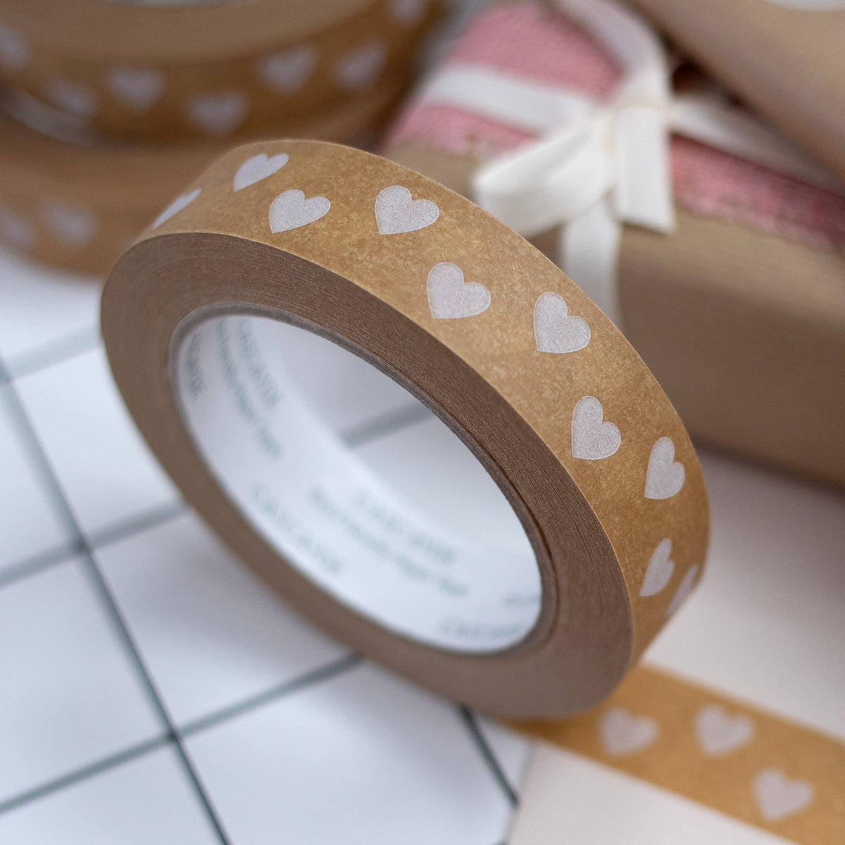 A roll of brown paper packing tape with a repeating white heart pattern is shown in a styled flat-lay setting alongside wrapped gift boxes and a pink card.