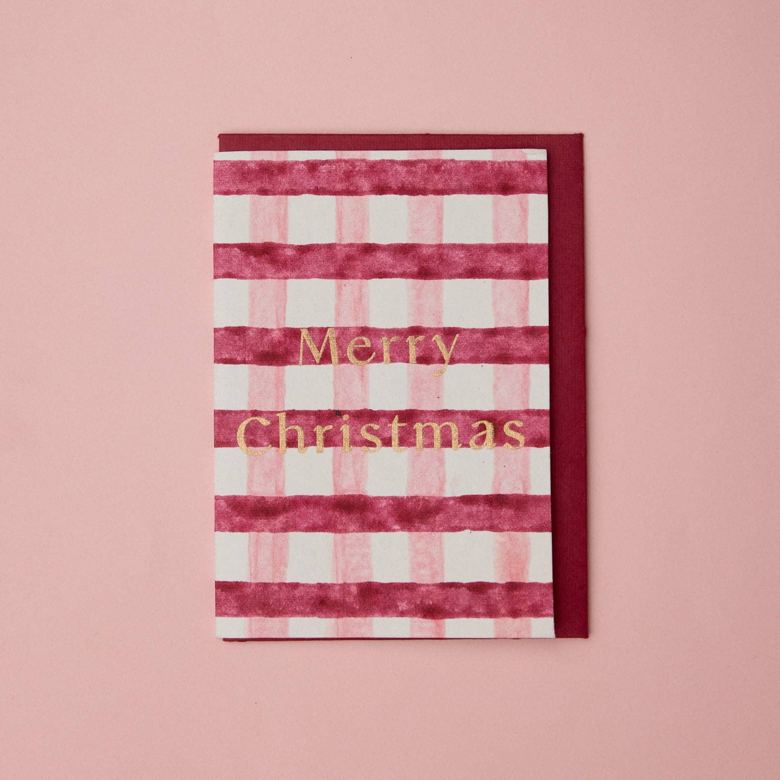 A Christmas greeting card with horizontal pink and white stripes and gold foil text reading 'Merry Christmas,' shown with a matching dark red envelope on a soft pink background.