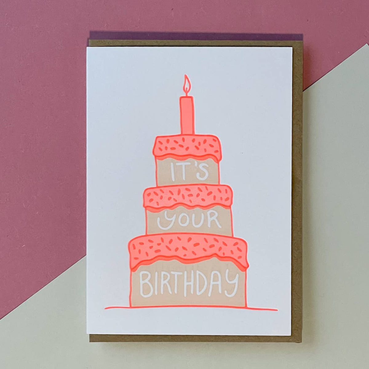 An A6 birthday greeting card with a brown envelope, featuring an illustration of a three-tier cake with a candle and the words 'IT'S YOUR BIRTHDAY' on a white card, set against a pink and cream background.