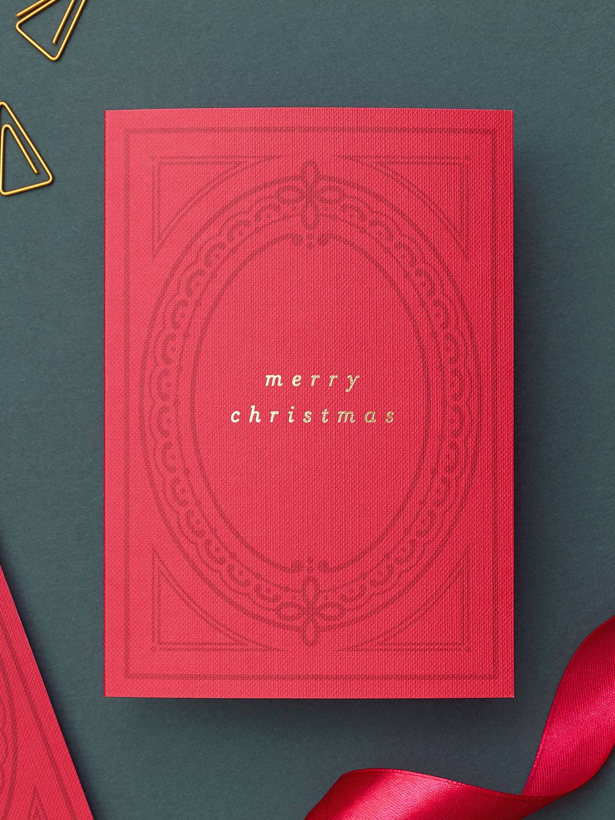 A red textured Christmas greeting card with embossed decorative borders and the words 'merry christmas' in gold foil, displayed on a dark green background with a red ribbon and gold paperclip props.
