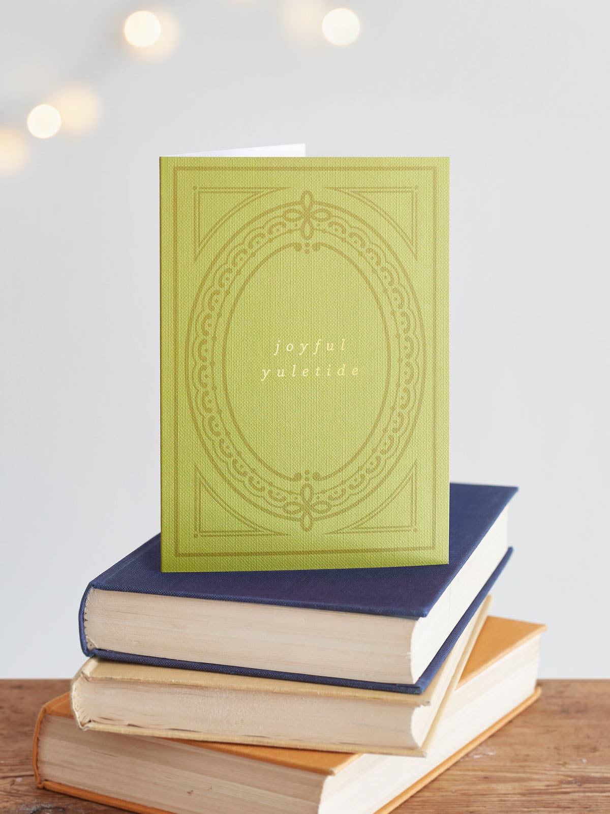 A yellow-green christmas card with the words 'joyful yuletide' with an embossed oval border design, standing upright on a stack of three other books on a wooden surface against a softly blurred light gray background with warm lights.
