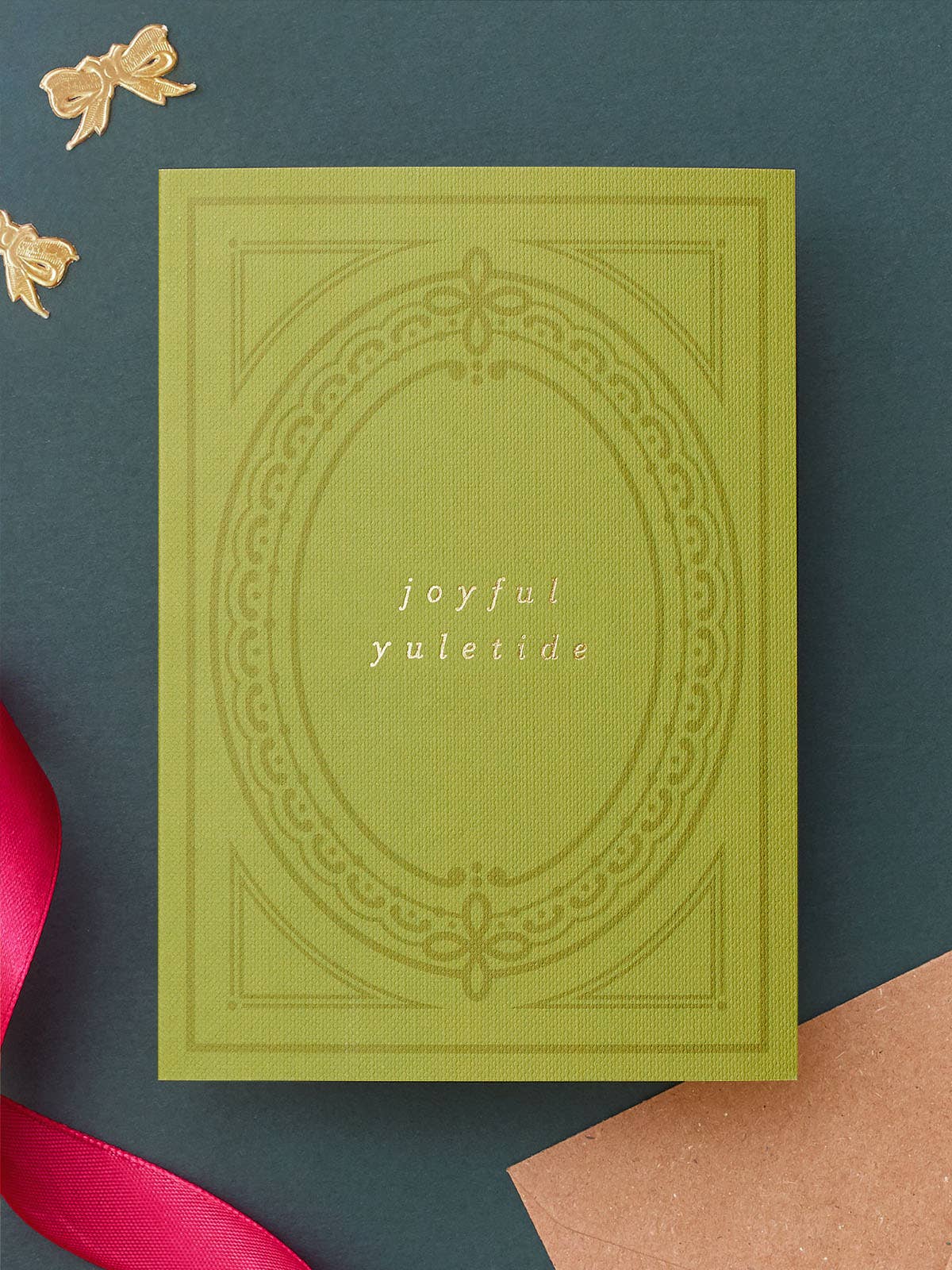 An A6 olive green greeting card with embossed decorative border details and the words 'joyful yuletide' in the centre, styled on a dark teal background with ribbon and paper props.