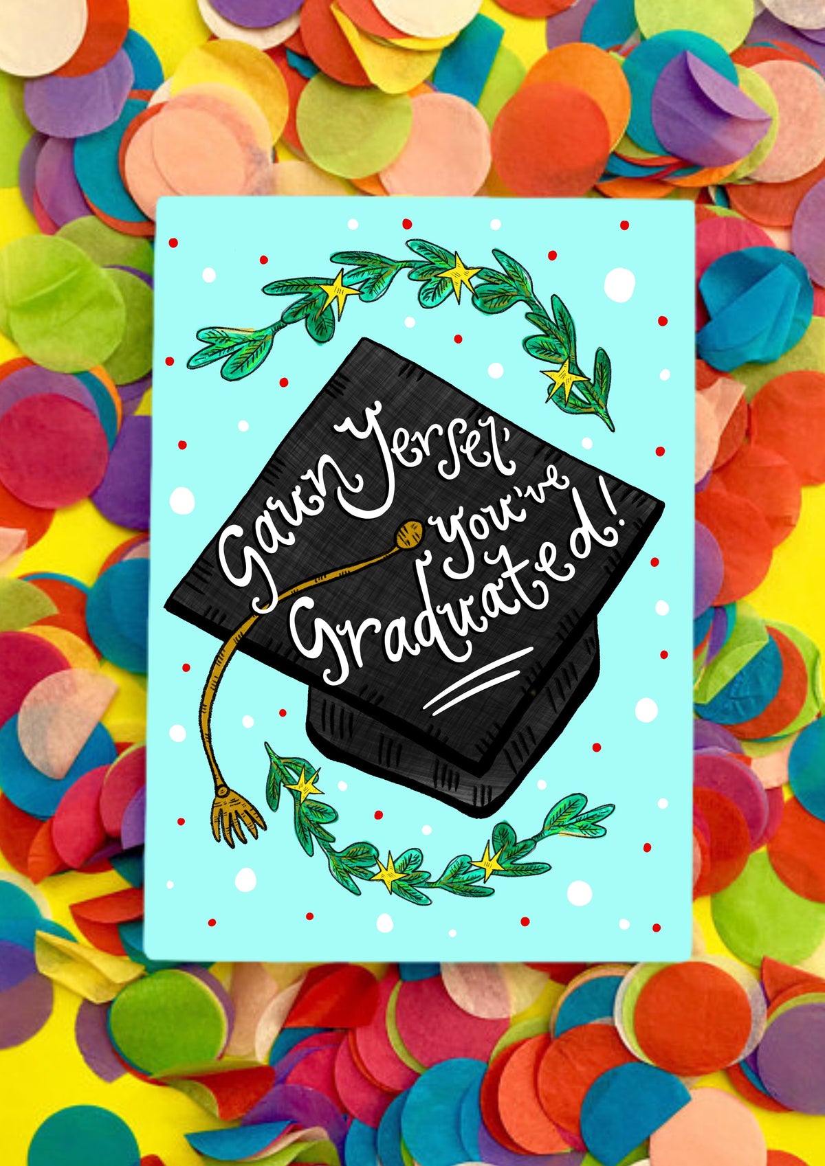 A greetings card wiht a light turquoise background and a large graduation black cap in the middle with gold tassel. In white writing, it says on the cap 'Gaun Yersel' you've graduated!' in script handwriting. There are leafy branches at the bottom and top of the card also. The card is sitting on a bed of rainbow confetti.