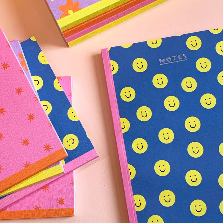 A close up image of notebooks, the main one is navy and covered in yellow happy faces and says NOTES in silver foil at the top.