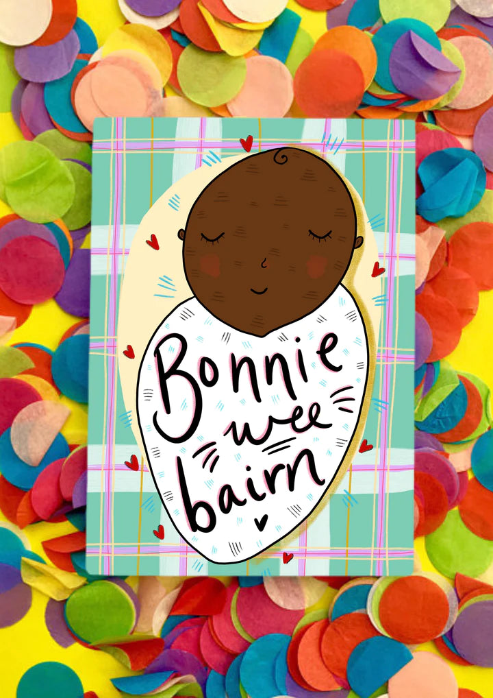 A greetings card with green and pink tartan background and Afro heritage baby swaddled in a white blanket, with wording Bonnie Wee Bairn across it.