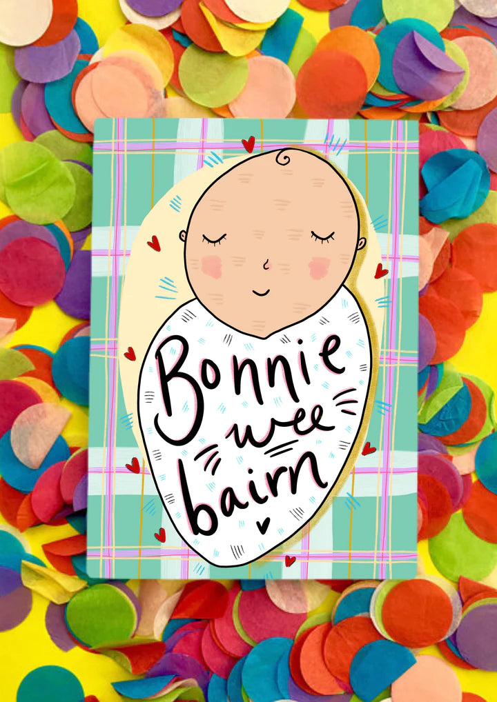 A greetings card with green and pink tartan background and a white baby swaddled in a white blanket, with wording Bonnie Wee Bairn across it.